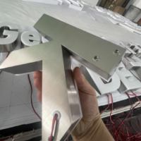 Hand houdt de achterkant van een metalen doosletter met geborsteld RVS, met zichtbare bedrading en montagepunten op een werktafel met andere letters op de achtergrond.”