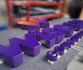 Paarse 3D doosletters met de tekst ‘WiseUp Consultancy’ liggen op een werktafel in een werkplaats. De grote letters ‘WiseUp’ staan op de voorgrond en hebben een paarse kleur met een oranje verloop op de letter ‘U’. De kleinere letters ‘Consultancy’ liggen eronder. De achtergrond met gereedschap en een rode werkbank is onscherp (blurred), waardoor de focus op de glanzende letters ligt.