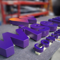 Paarse 3D doosletters met de tekst ‘WiseUp Consultancy’ liggen op een werktafel in een werkplaats. De grote letters ‘WiseUp’ staan op de voorgrond en hebben een paarse kleur met een oranje verloop op de letter ‘U’. De kleinere letters ‘Consultancy’ liggen eronder. De achtergrond met gereedschap en een rode werkbank is onscherp (blurred), waardoor de focus op de glanzende letters ligt.