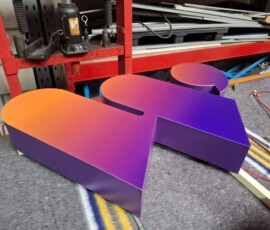 Grote 3D letter ‘W’ met een kleurverloop van oranje naar paars, liggend op een werktafel in een werkplaats. De doosletter heeft een strakke, matte afwerking en ligt op afstandhouders klaar voor montage, met gereedschap en een rode werkbank op de achtergrond.